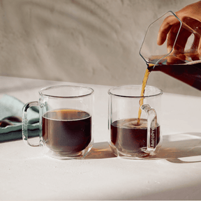 Person pouring coffee from a clear glass carafe into two clear glass mugs on a light surface.