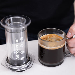 AeroPress Coffee Maker - Clear next to mug with coffee  #color_clear