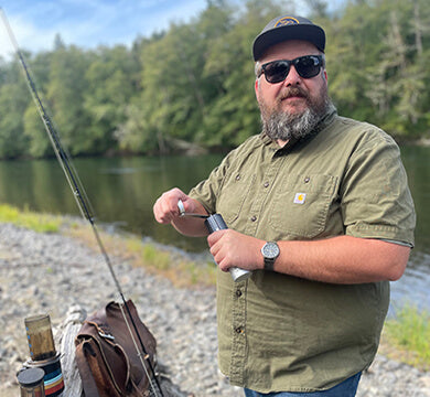 Man using a manual coffee grinder by a river with trees in the background