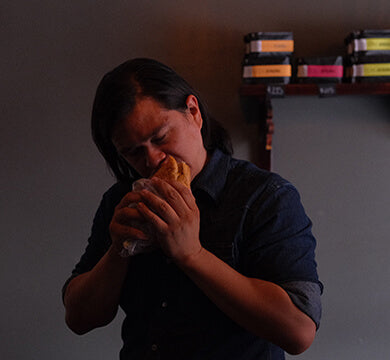 Person eating a sandwich in a dimly lit room with shelves in the background