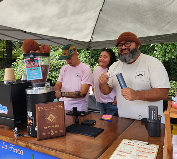 Three people at a coffee booth with coffee equipment and a 'First Place' award.