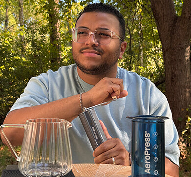 Man holding a manual coffee grinder with an AeroPress coffee maker outdoors with trees in the background