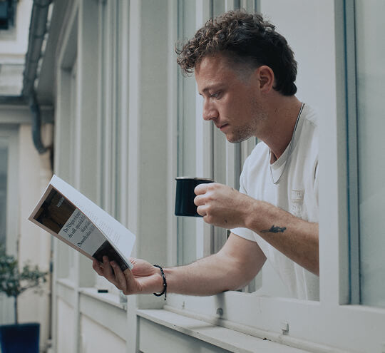 Man reading a book and holding a mug by a window
