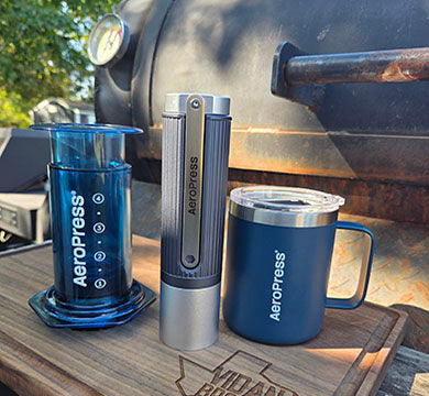 AeroPress coffee maker, manual coffee grinder and mug on a wooden surface outdoors