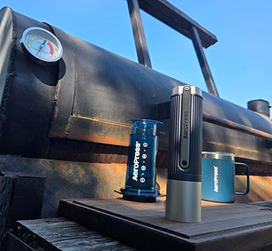 Manual coffee grinder, AeroPress coffee maker and mug next to a barbeque smoker