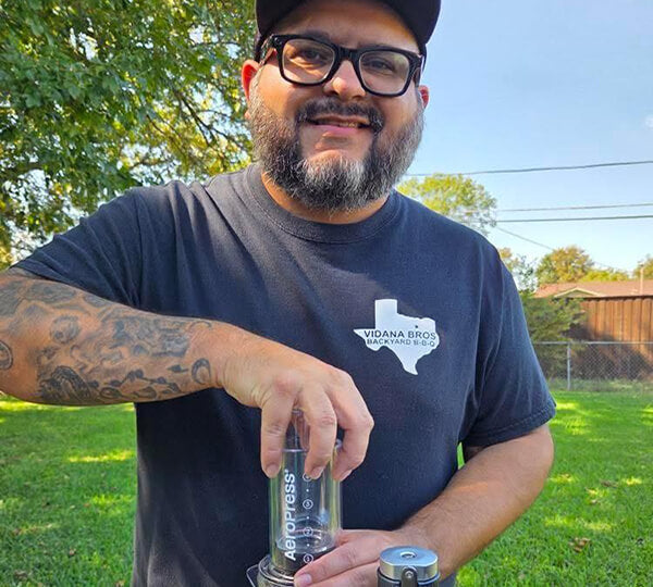 Man brewing with an AeroPress coffee maker outdoors with trees and grass in the background