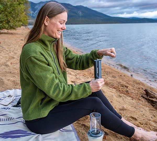 Woman sitting by a lake, using a portable coffee grinder.