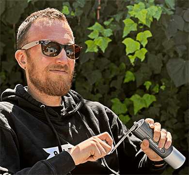 Man wearing sunglasses and a black hoodie holding a manual coffee grinder against a green leafy background