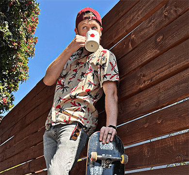 Person drinking from a coffee cup while holding a skateboard against a wooden wall.