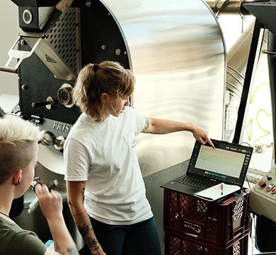 Person operating a coffee roaster with another person observing, in a coffee roastery setting.