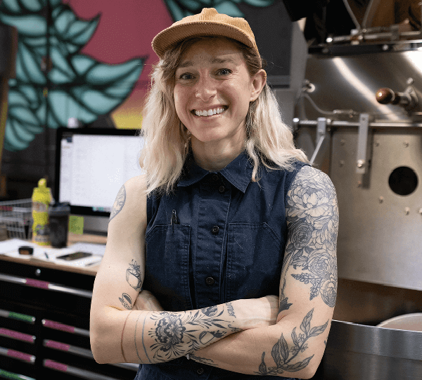 Person with tattoos wearing a cap and denim vest in a coffee roastery setting