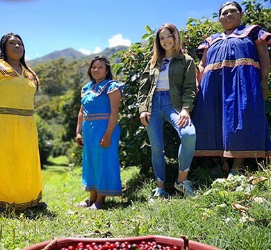 Four women, three in traditional attire and one in modern clothing, standing outdoors with a scenic background.