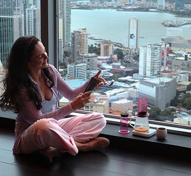 Woman sitting by a large window with a cityscape view, using a manual coffee grinder.