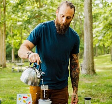 Man with tattoos outdoors, holding a kettle and a coffee press, surrounded by greenery.