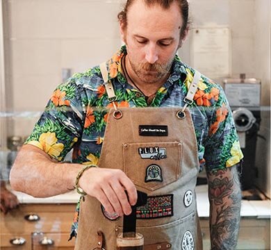 Man wearing a floral shirt and brown apron with coffee shop branding, standing in a kitchen.