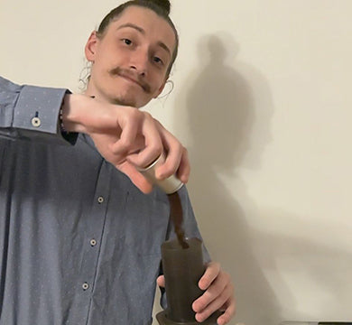 Person pouring ground coffee into an AeroPress coffee maker against a white wall
