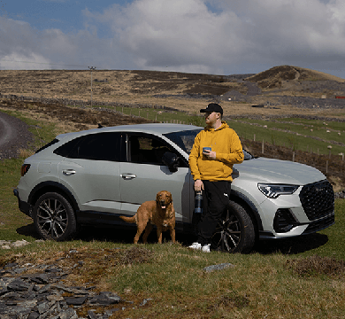 Person in yellow jacket and cap standing next to a silver SUV with a dog on a grassy road.