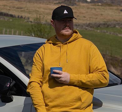 Man in yellow hoodie and black cap holding a blue cup, standing next to a car in a rural setting.