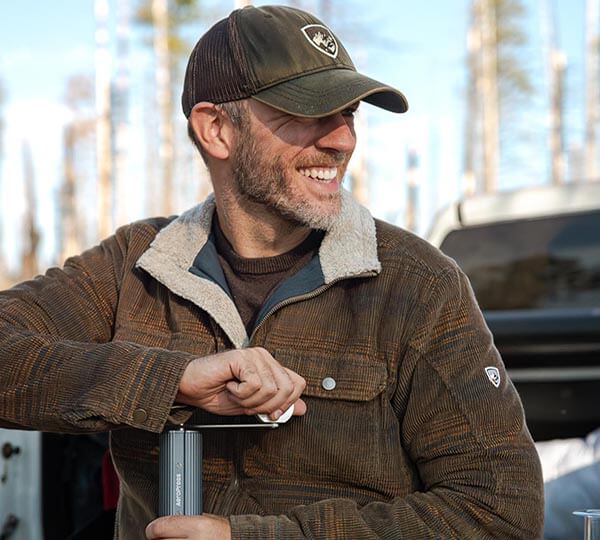 Man wearing a brown cap and jacket with a logo, standing outdoors with trees in the background.