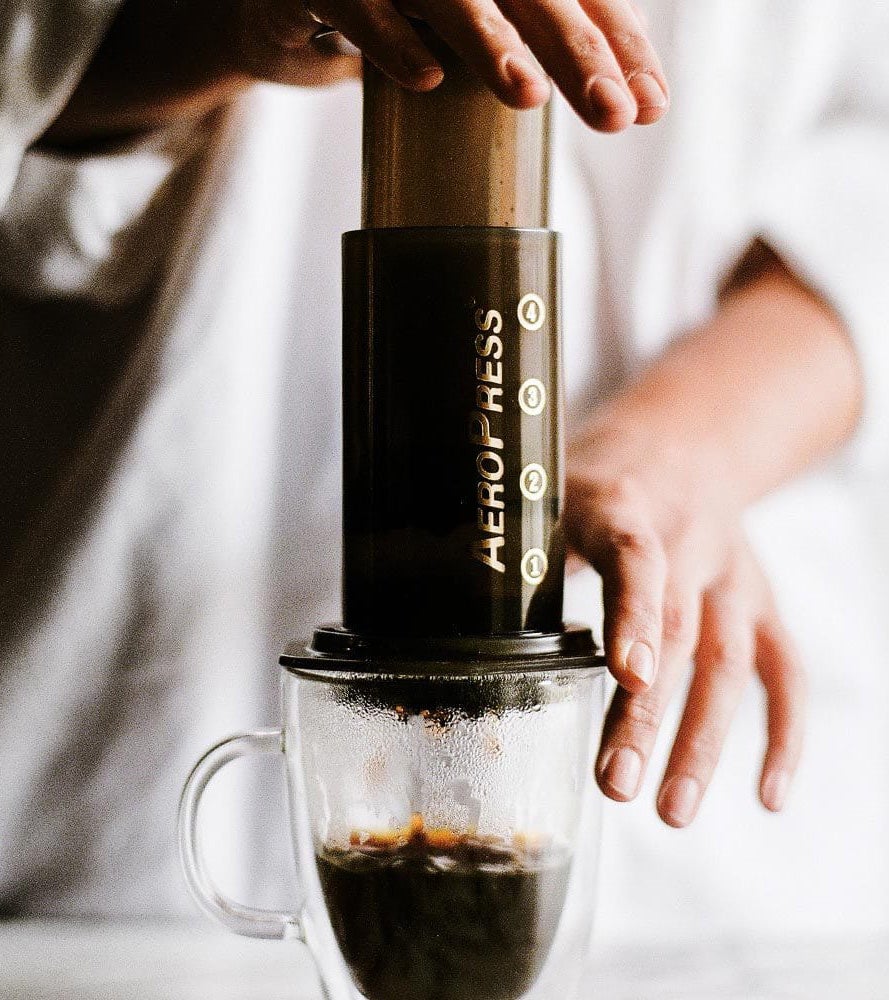 Ein Mann presst eine AeroPress aus Holz und Glas über eine Tasse, daneben liegt sein Rühr-Löffel – Ansicht 2
