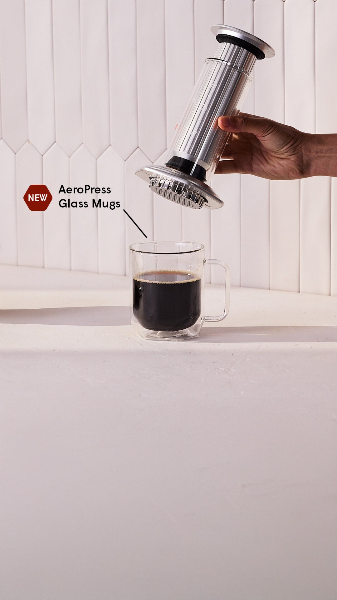 Person using an AeroPress Premium to make coffee in a glass mug on a white surface.