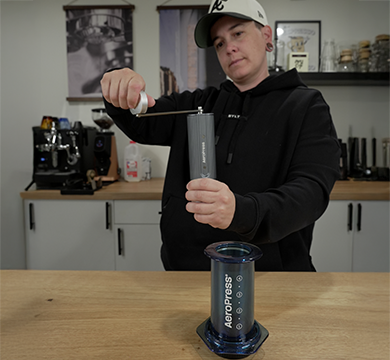 Person using an AeroPress coffee maker and manual coffee grinder in a kitchen setting