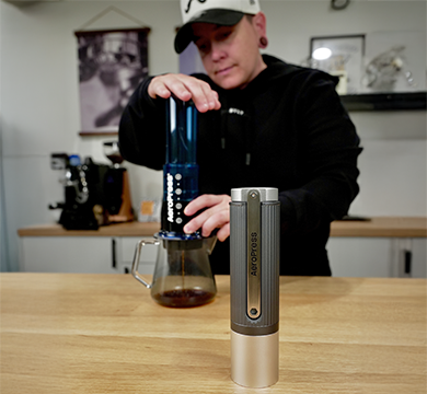 Person brewing AeroPress coffee next to a manual coffee grinder
